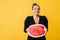 Pretty laughing girl in black dress happily looking aside while holding half of big watermelon in hands over yellow