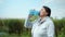 Pretty old woman quenches thirst drinks water after doing sports outdoors in meadow by river