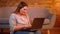 Pretty overweight student sits on floor typing smilingly on laptop being satisfied with job in cozy home atmosphere.