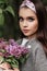 Pretty portrait of woman in bandana in fashionable gray coat with color make-up with bouquet chic purple flowers lilac near green
