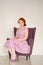 Pretty redheaded pin up woman wearing pink polka dot dress and posing with purple armchair on white background