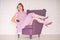 Pretty redheaded pin up woman wearing pink polka dot dress and posing with purple armchair on white background