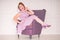 Pretty redheaded pin up woman wearing pink polka dot dress and posing with purple armchair on white background