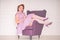 Pretty redheaded pin up woman wearing pink polka dot dress and posing with purple armchair on white background