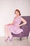 Pretty redheaded pin up woman wearing pink polka dot dress and posing with purple armchair on white background