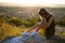 Pretty sad woman in black short summer dress sitting on a rock thinking outdoors at sunset. Fashionable female contemplating in