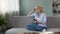 Pretty senior female sitting on sofa and calculating budget, using smartphone