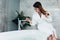 Pretty slim woman wearing bathrobe sitting on edge of bathtub filling up with water