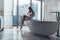 Pretty slim woman wearing bathrobe sitting on edge of bathtub filling up with water