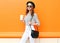 Pretty smiling woman with coffee cup wearing fashion black hat white pants handbag clutch over colorful orange