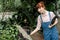 Pretty smiling woman gardener inspecting the quality of plants in modern greenhouse, holding a clipboard with notes and