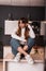 Pretty stylish young woman with red hair in trendy casual blue-white clothes posing sitting on a wooden table in the cook room.