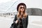 Pretty urban hipster young woman in a fashionable hat in stylish sunglasses in a vintage leather jacket with a backpack walks