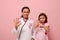 Pretty woman doctor in medical gown and a cute little girl, both with pink ribbon and phonendoscope around their neck, gesticulate