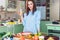 Pretty woman holding a glass of juice, smiling, looking at camera, standing table with fresh fruit and vegetables o