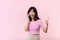 Pretty young asian woman showing success, victory hand gesture while receiving great news from smartphone on pink background.