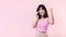 Pretty young asian woman showing success, victory hand gesture while receiving great news from smartphone on pink background.