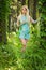 Pretty young blonde girl with closed eyes and long hair in turquoise dress standing in the green forest where trees are enlaced wi
