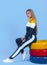 Pretty young blonde model girl in sportswear posing next to car multi-colored tires in blue backround