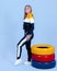 Pretty young blonde model girl in sportswear posing next to car multi-colored tires in blue backround