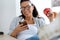 Pretty young business woman using her mobile phone while eating red apple in the office