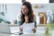 Pretty young business woman working with computer while consulting some invoices and documents in the kitchen at home