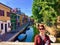 A pretty young female tourist posing on a bridge for a photo with the old colourful historic homes and canal of Burano, Italy