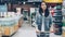 Pretty young lady is walking through aisle in supermarket with shopping cart looking at shelves with products, employees