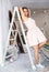 Pretty young landlady in beige dress poses playfully on stepladder in renovated cottage