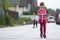 Pretty young long-haired blond child girl in pink clothing with scooter on sunny suburb street and boy on skateboard blurred