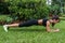 Pretty young sportswoman doing plank exercise on grass in city park