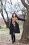 Pretty young woman in black coat and colorful scarf stand in park by tree full body shot winter