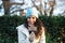 pretty young woman with curly brown hair wears a woollen hat for the cold and covers her face with a dry leaf from a tree. Autumn