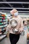 Pretty young woman inmask, pink knitted hat and sweater poses humorously in supermarket with two colanders in kitchen utensils