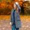 Pretty young woman model in gray trendy coat in a vintage blouse with a handbag stands in a park on the background of autumn trees