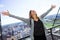 Pretty young woman with raised arms to the sky enjoying the time at modern rooftop of high-rise building.