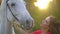 Pretty young woman in red dress caressing graceful white horse at sunset. Harness, Muzzle horse, mane. Woman`s hand