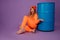 Pretty young woman sitting near blue barrel on stylish orange overall and hat on isolated over violet background.