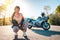 Pretty young woman with tattoos on her arm, wearing leather jacket, and posing sitting. Road and motorbike in the