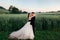 Prim newlyweds stand hugging before the field with green wheat