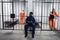 A prison warden guards cells and surfs the Internet via smartphone with dangerous prisoners in orange prison uniforms.