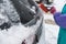 Process of cleaning a car from snow in the morning, girl removing snow from windscreen with a window scraper brush after snowstorm