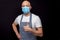 Professional bald butcher or fishmonger in white t-shirt and black and white apron on dark background. Man wearing blue face mask