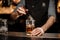 Professional bartender adding a big ice cube to the measuring glass cup with a brown cocktail