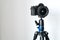 Professional black mirrorless camera stands on a tripod against a white wall.
