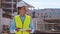 Professional builder standing in front of construction site. Foreman in hardhat helmet and vest. Office building and