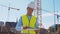 Professional builder standing in front of construction site. Foreman in hardhat helmet and vest. Office building and