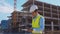 Professional builder standing in front of construction site. Foreman in hardhat helmet and vest. Office building and