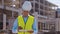 Professional builder standing in front of construction site. Foreman in hardhat helmet and vest. Office building and