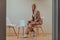 A professional businesswoman sits on a chair, surrounded by a serene beige background, diligently working on her laptop